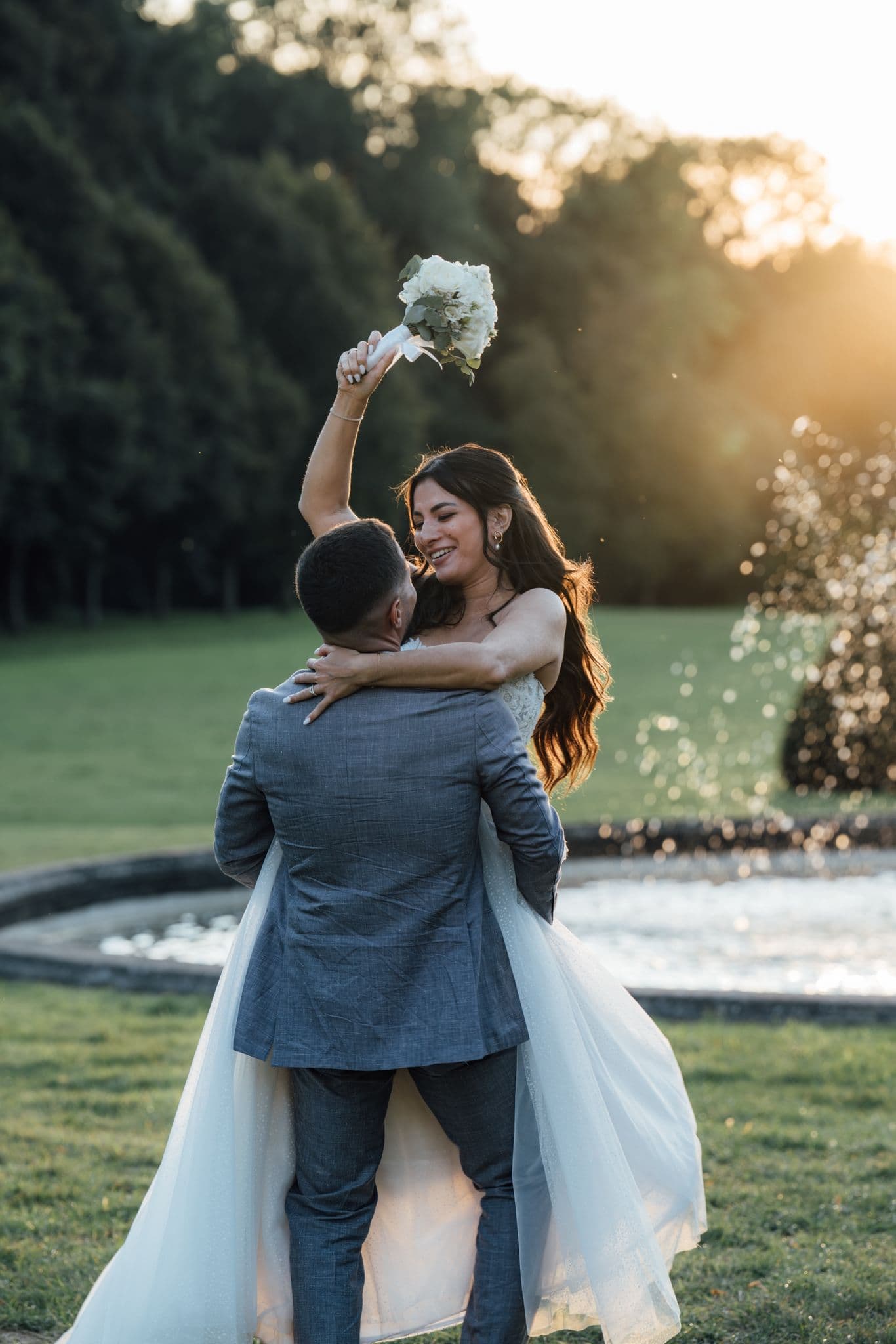 mariés marine et maxime posant et levant son bouquet de fleurs devant la fontaine du sublime château de champlatreux